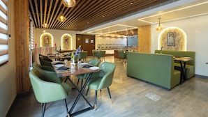 Un restaurant moderne avec un intérieur caractérisé par des poutres en bois au plafond, des banquettes recouvertes d’un tissu vert et un espace de restauration central doté de tables et de chaises.