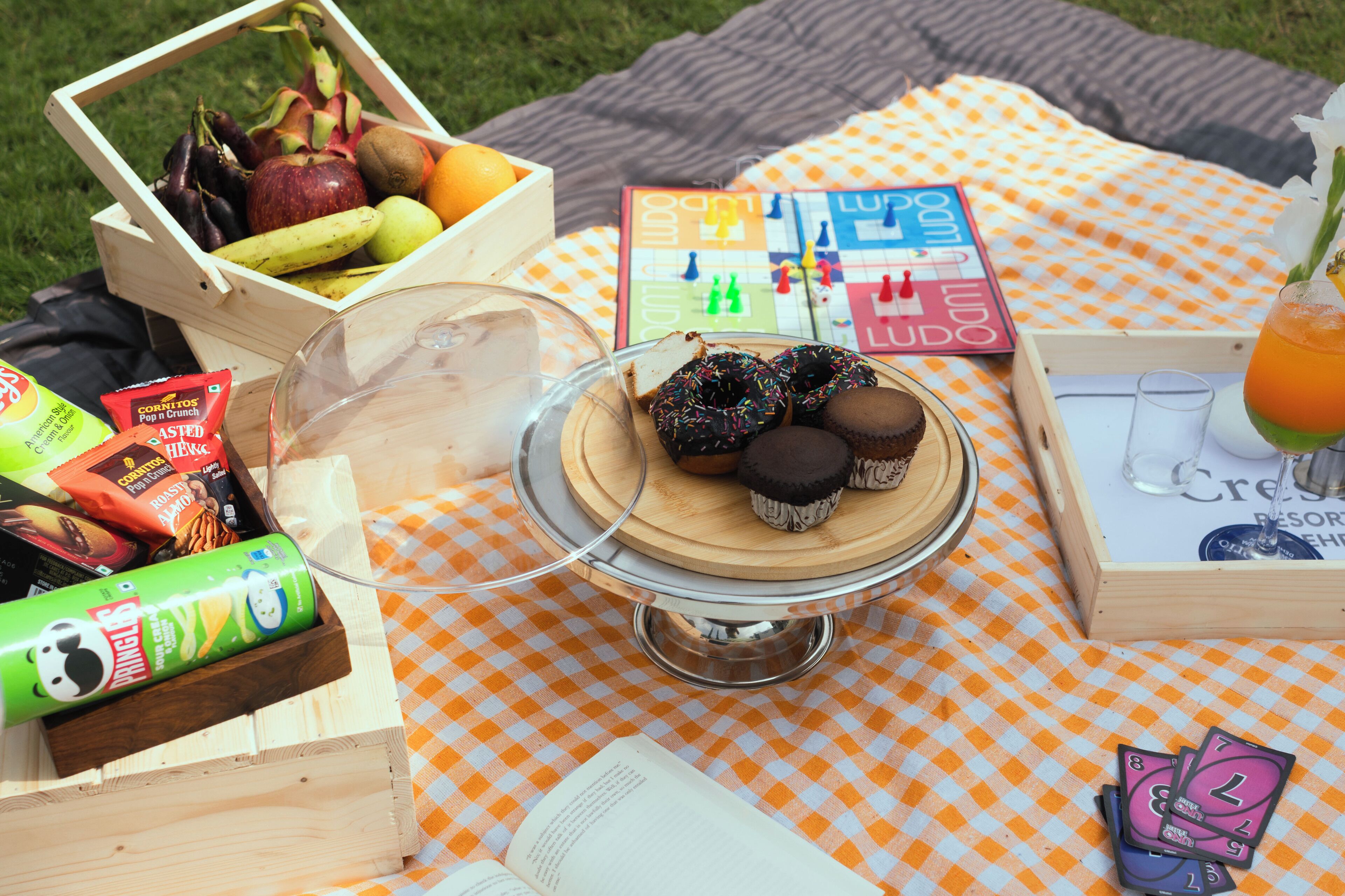 Un montaje para picnic con una manta a cuadros, un cesto de madera con frutas, un vaso de jugo y una tabla con cupcakes.