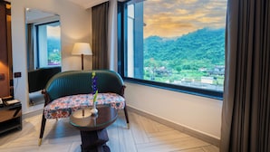 Une chambre d’hôtel avec une grande fenêtre, un fauteuil recouvert d’un tissu vert, une petite table avec un vase et une lampe.