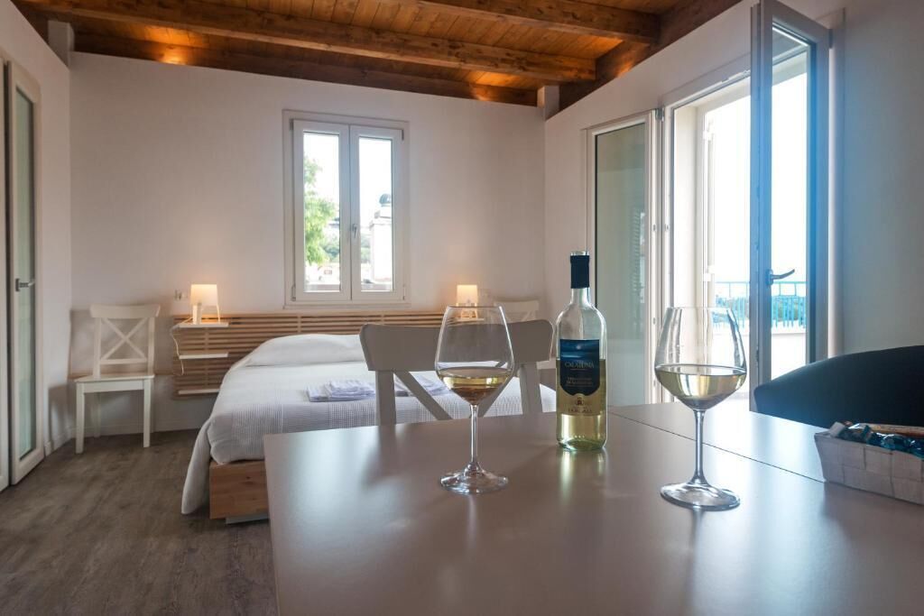 Une chambre d’hôtel avec un lit, une table avec deux verres à vin et une bouteille, une chaise et une fenêtre donnant sur un paysage.