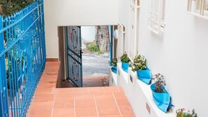 A narrow corridor with a blue metal fence on one side, a white wall with windows on the other, and terracotta-tiled flooring. - LOCANDA SU RECREU (Cala Gonone)