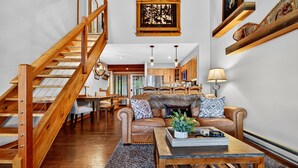 Un salón amplio con una escalera de madera, chimenea y una cocina al fondo.