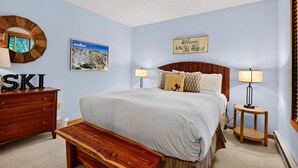 Un dormitorio con cama, cómoda de madera, banqueta, dos mesitas de noche con lámparas, un reloj de pared, un letrero de bienvenida y un cuadro enmarcado.
