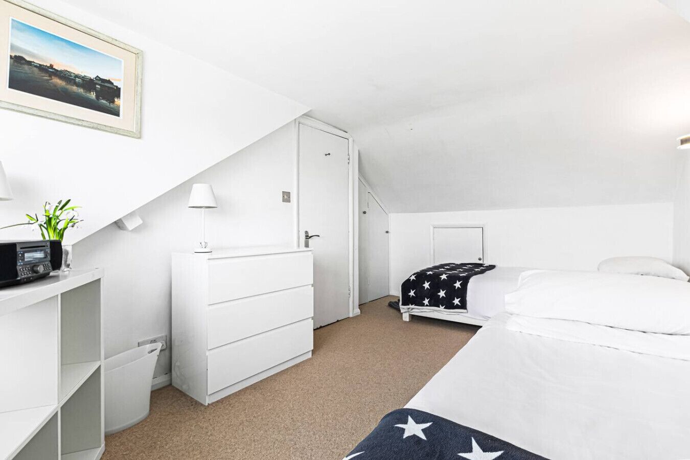 Une chambre à coucher blanche, avec un lit, une commode, un bureau avec une radio et un tableau accroché au mur.