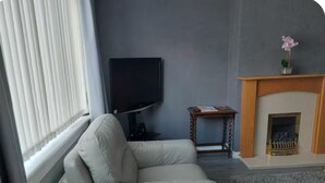 Ett vardagsrum med en vit soffa, en platt-TV, ett litet träbord och en öppen spis.