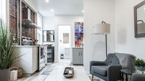 A modern kitchen with a brick wall, stainless steel appliances, and a white countertop. There is a dining area with a grey sofa and a coffee table. - The Hawthorne Apartment (Stratford)