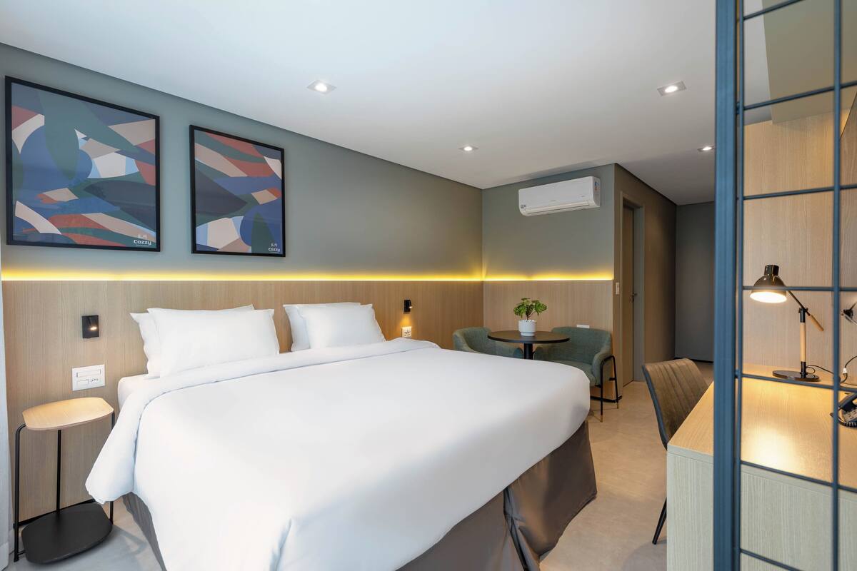 Habitación de hotel con cama, mesita de noche, silla, escritorio, lámpara y dos cuadros abstractos en la pared.