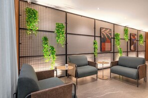 Un área de estar interior moderna con sillas y mesas acolchadas, una pared decorativa con plantas colgantes y obras de arte abstracto.
