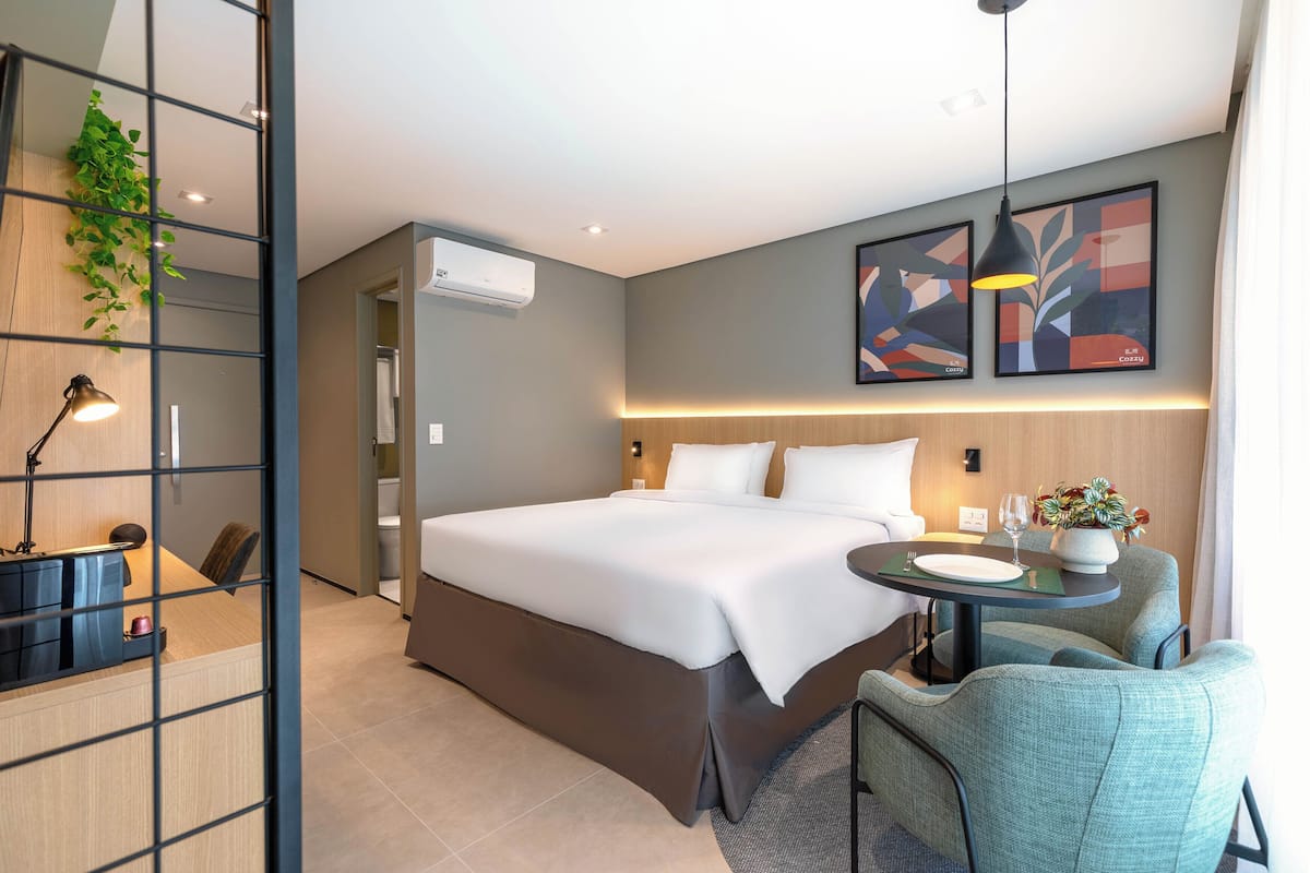 Una habitación de hotel moderna con una cama grande, un escritorio con una silla, una mesita con una planta y una obra de arte en la pared.