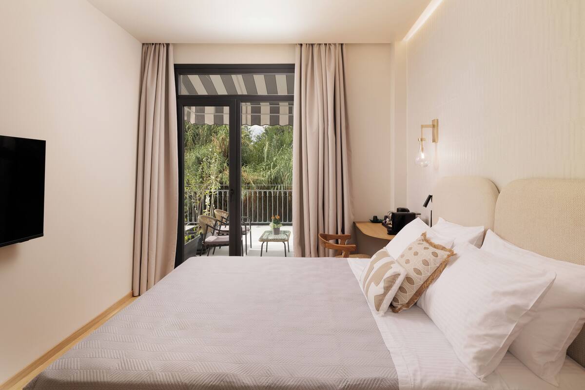 A hotel room with a large bed, a flat-screen TV, a balcony with a table and chairs, and a desk with a chair.