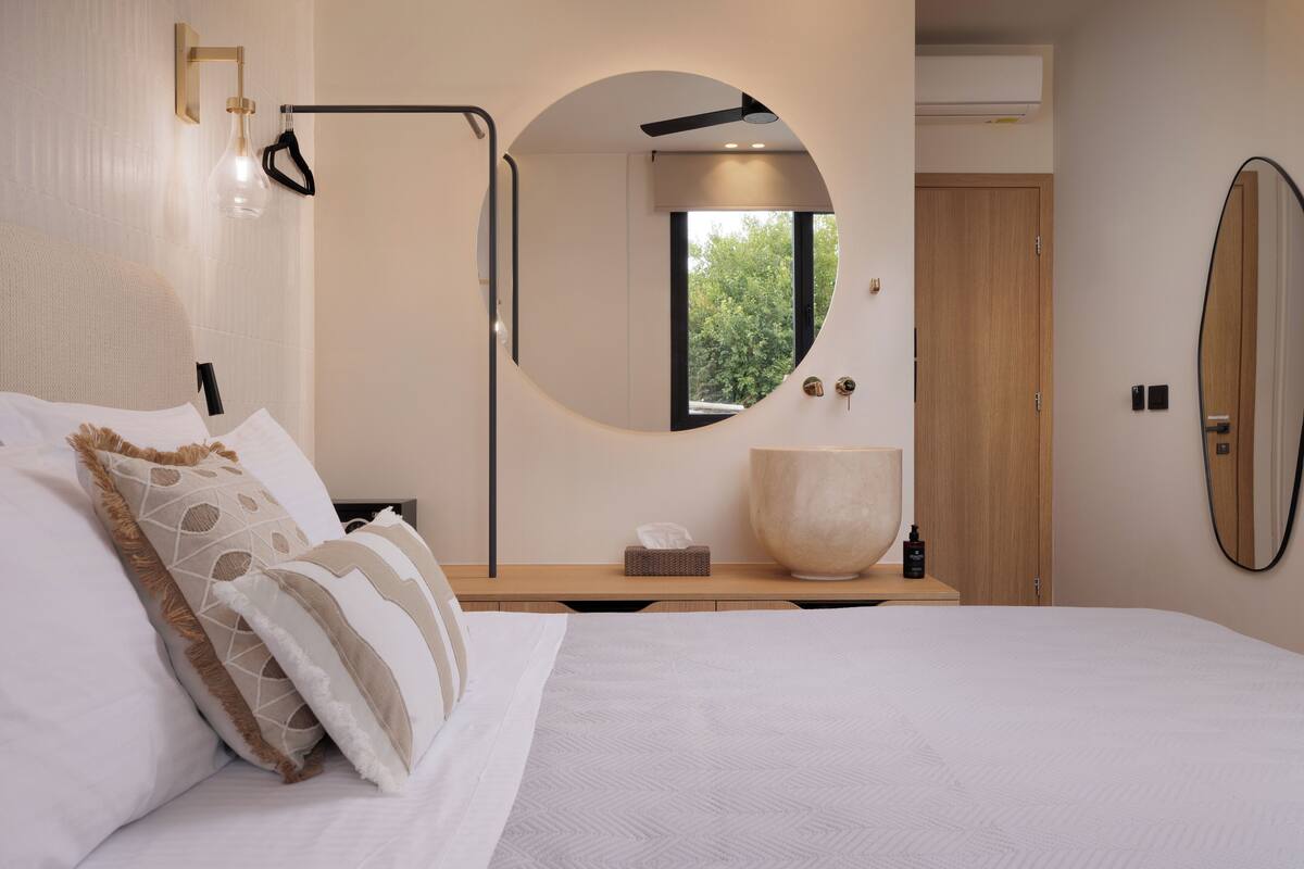 A modern bedroom with a large bed, a round mirror, a bedside table with a lamp, and a view of greenery through a window.