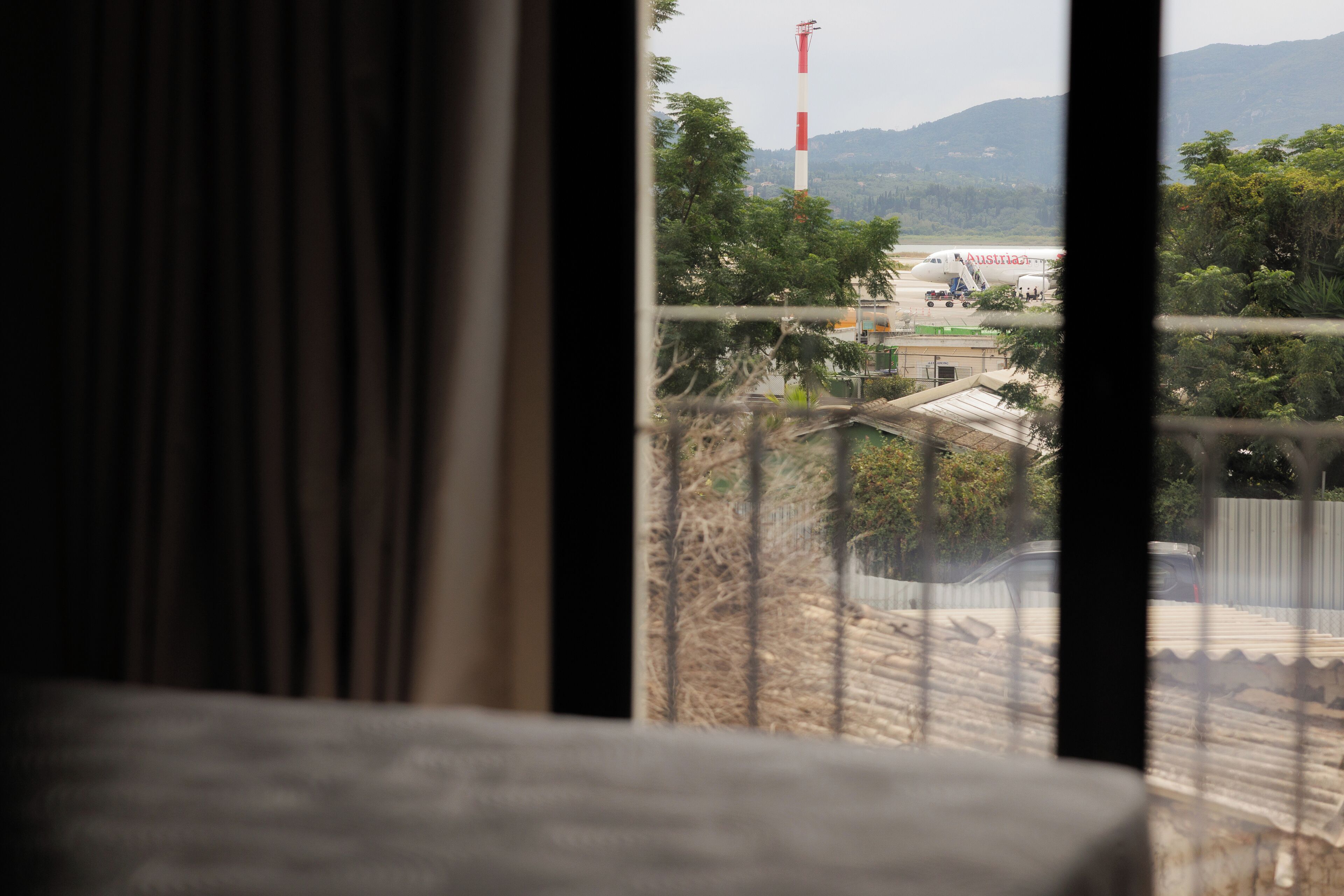 A view from a window with curtains, featuring an airport, a red and white tower, and a mountainous landscape.