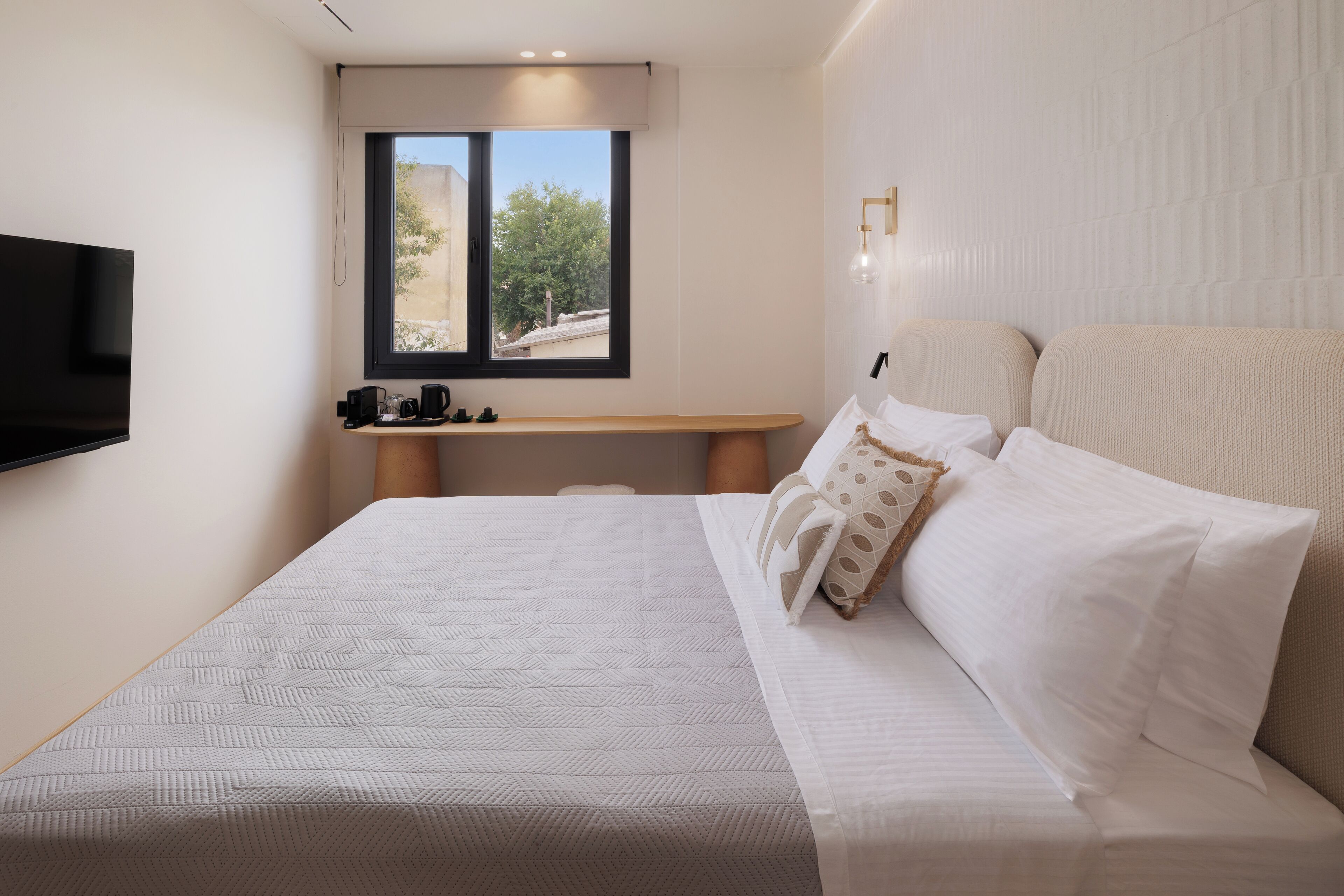A neatly arranged bedroom with a large bed, a bedside table, a flat-screen TV, and a window with a view of greenery.