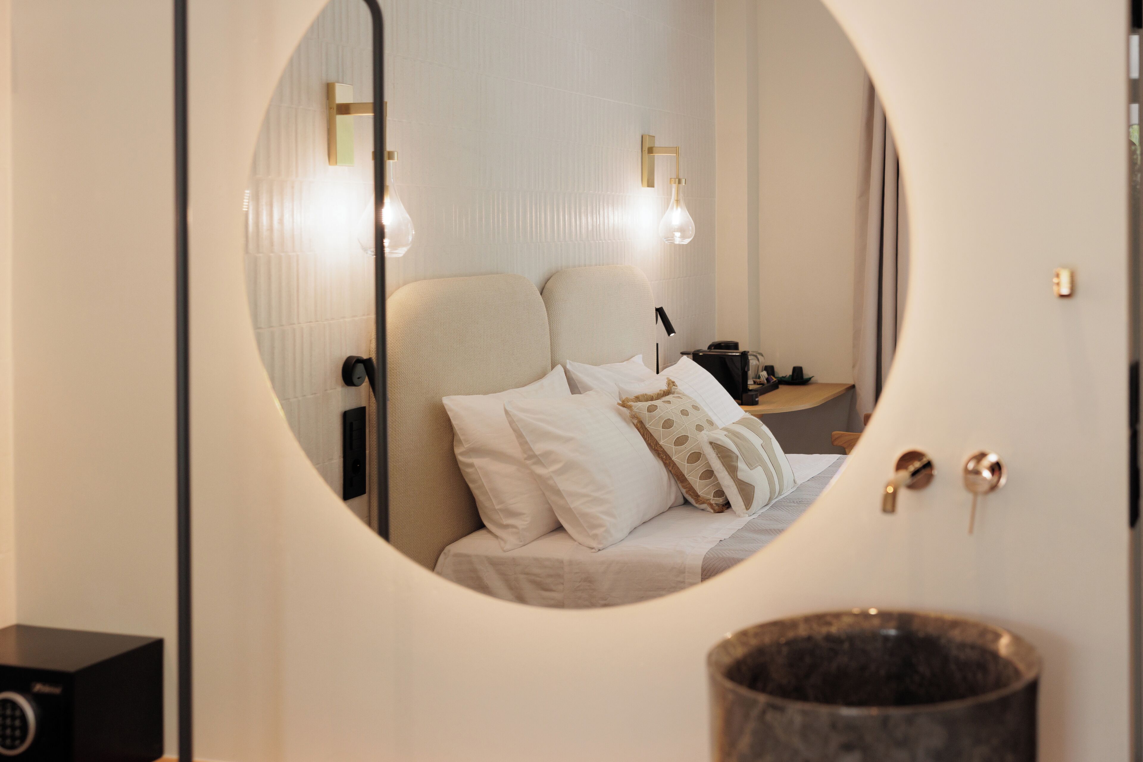 Un miroir rond reflète un lit avec une literie blanche et plusieurs oreillers, une table de chevet avec une lampe et une petite enceinte noire.
