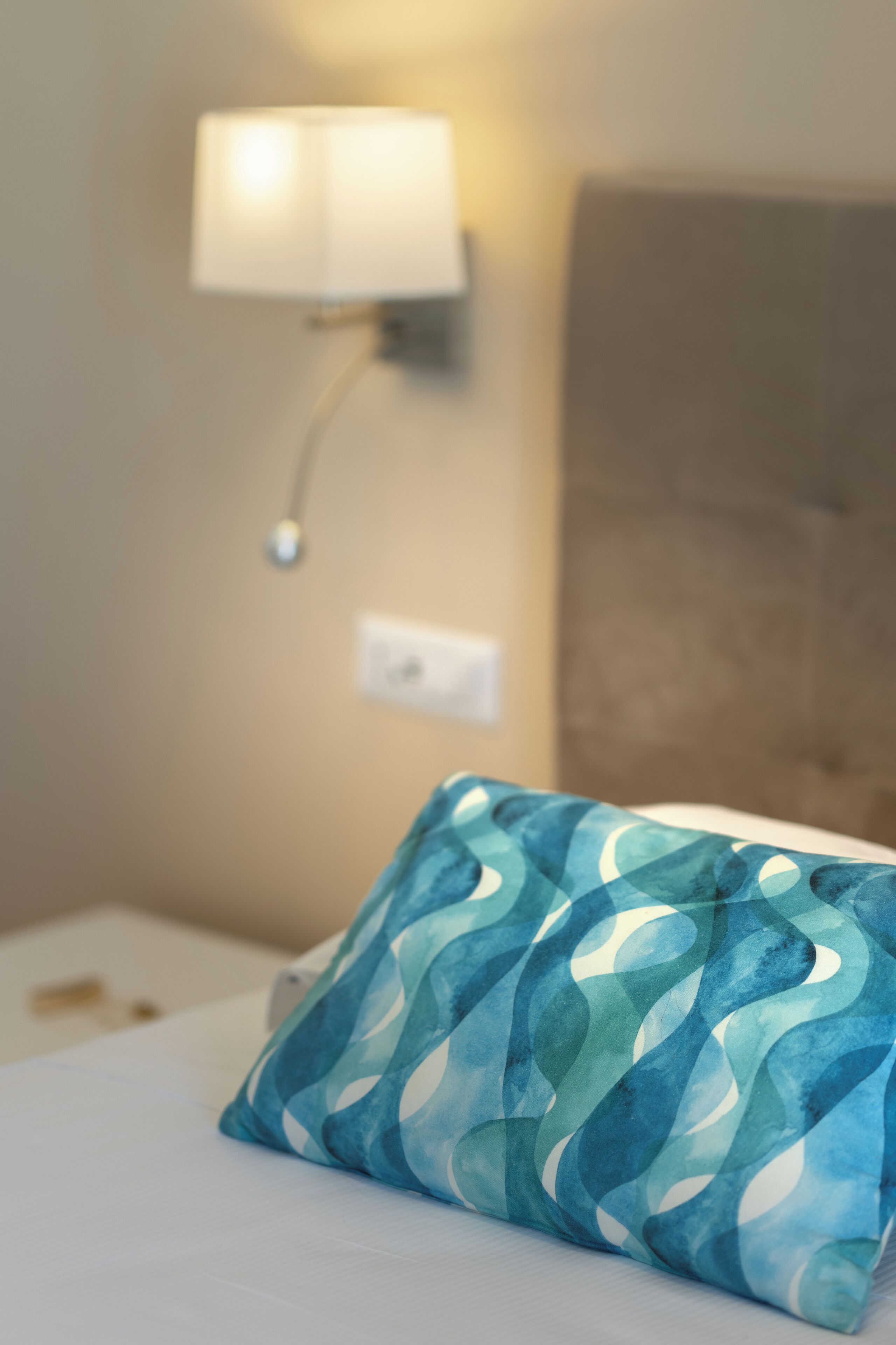 Un lit bien fait, agrémenté d’un coussin à motifs bleus, une lampe fixée au mur et un mur sobre.