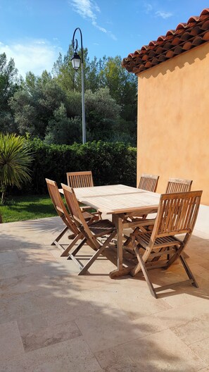 A wooden table and chairs set on a patio with a tiled floor, surrounded by greenery and a building with a tiled roof. - Villa at the foot of a hill (La Farlède)