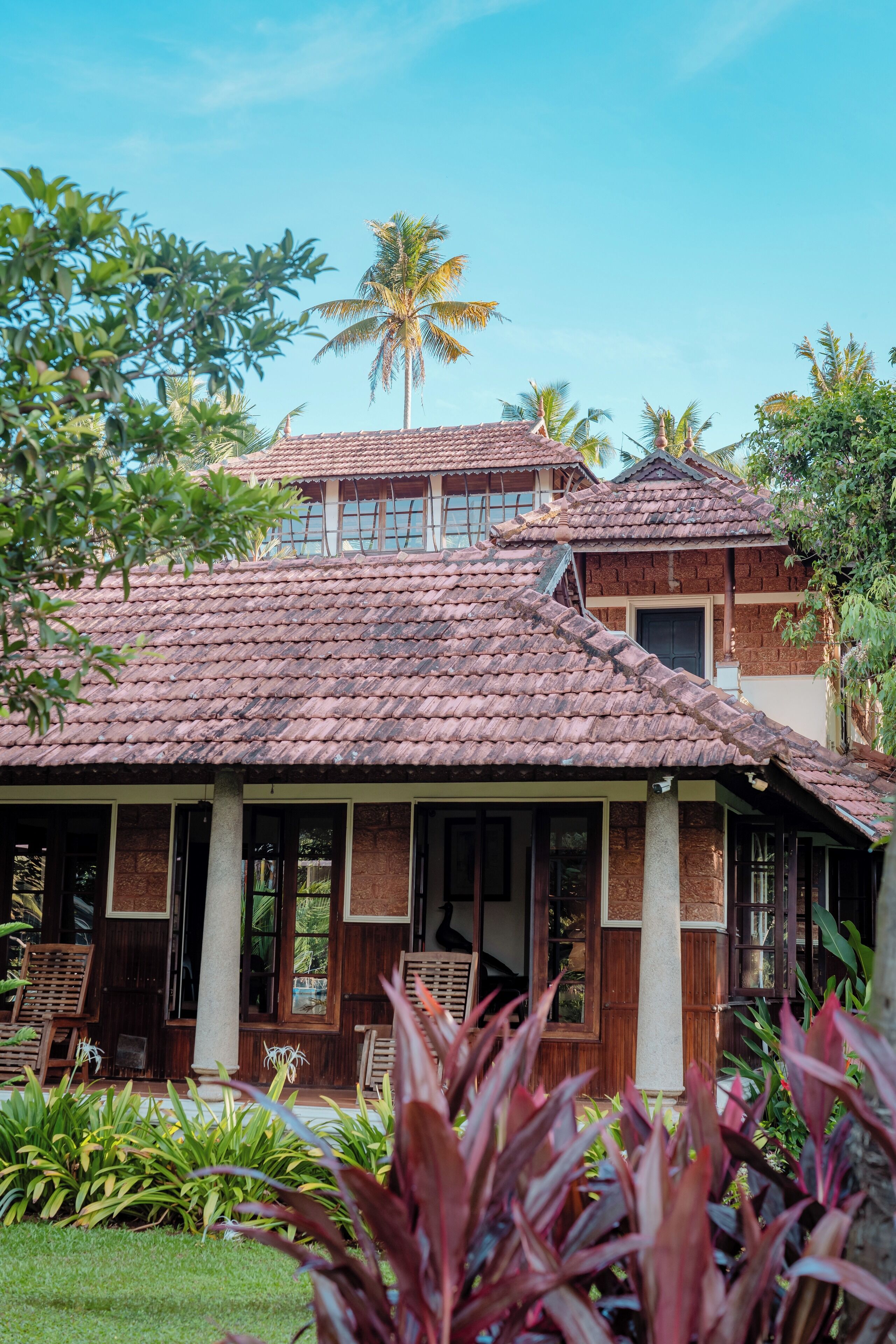 Une maison traditionnelle avec un toit en tuiles, des colonnes en bois et un jardin planté de plantes tropicales.