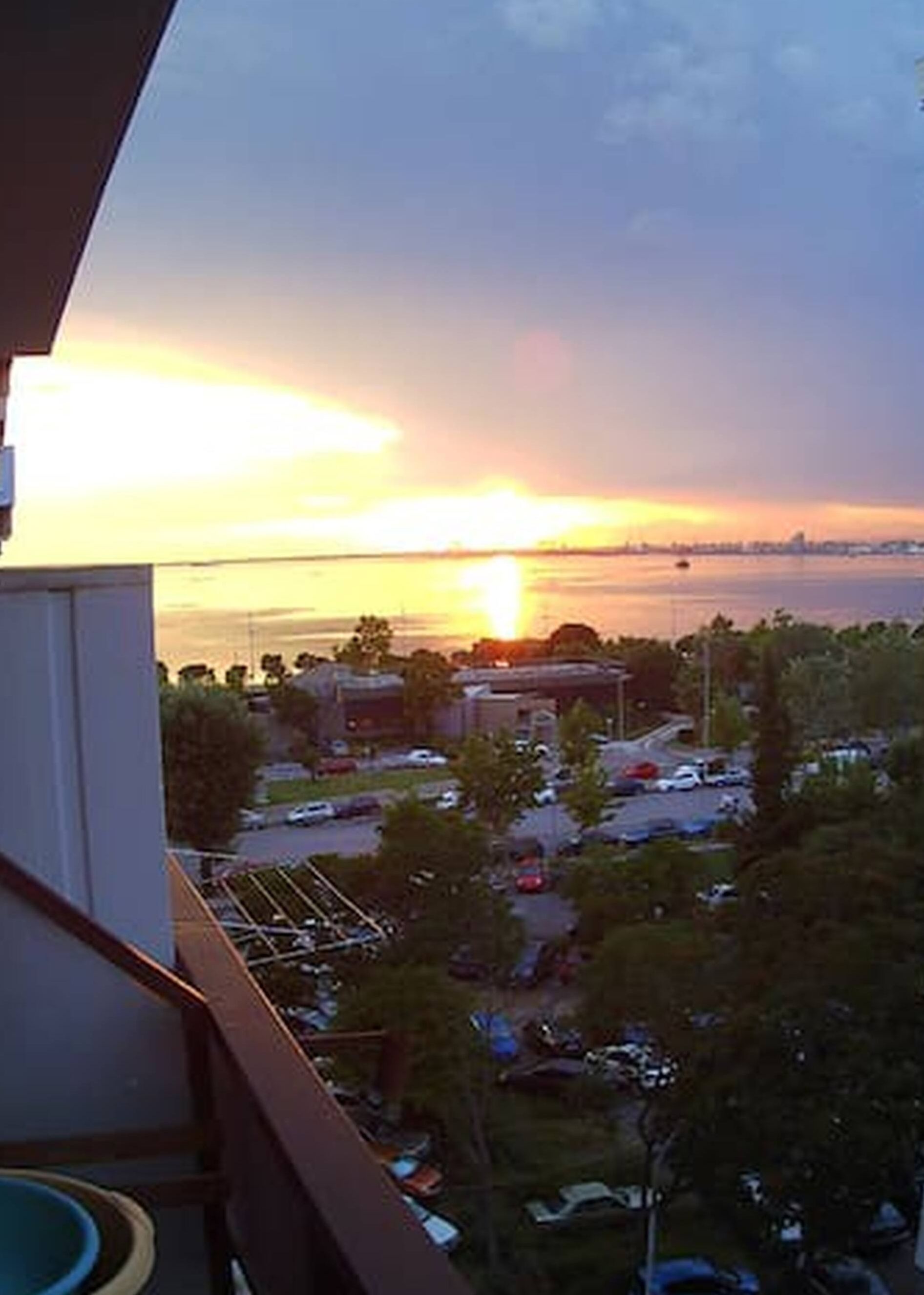 A view of a sunset over a body of water with buildings and parked cars in the foreground.