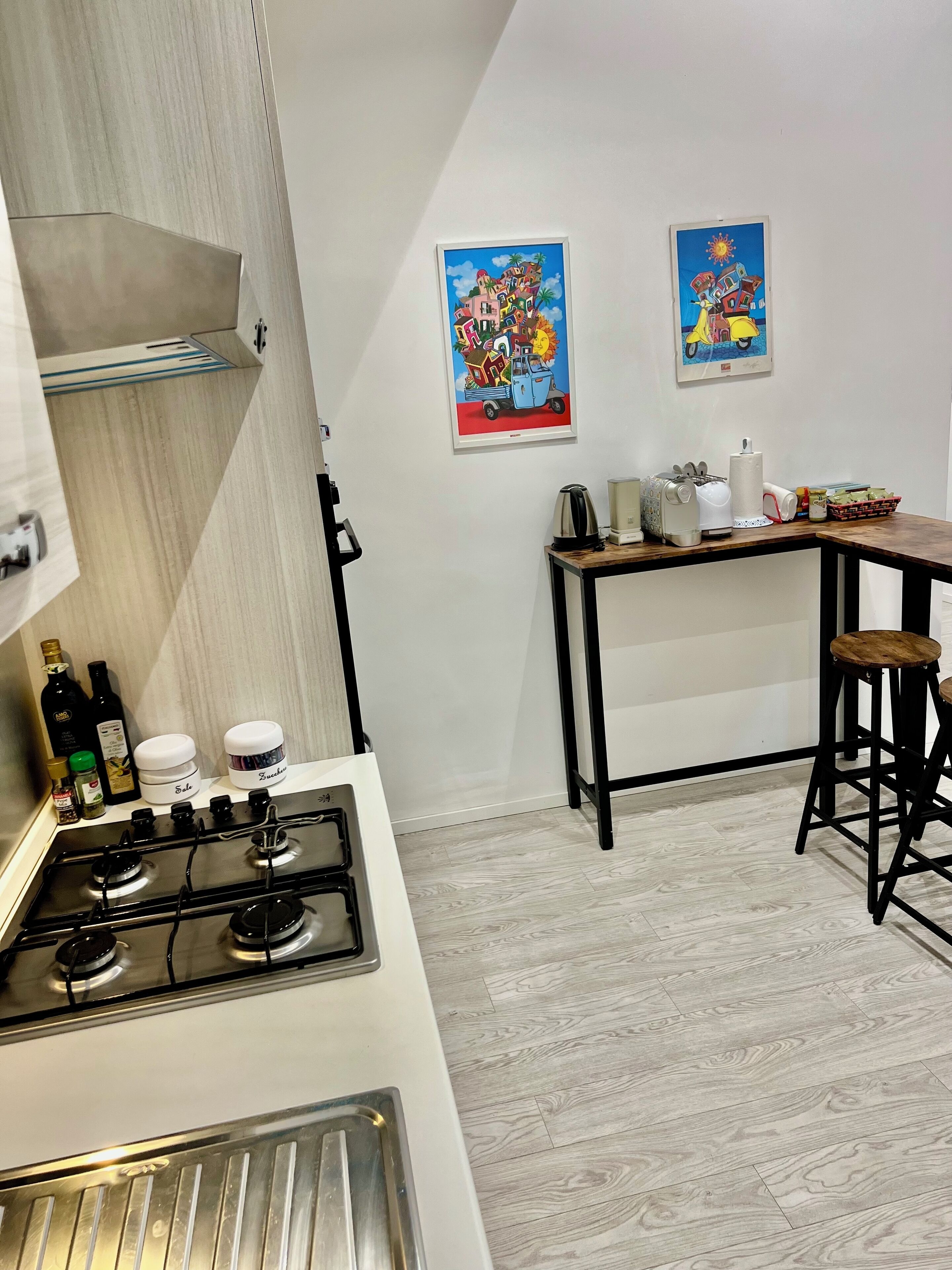Una cocina con estufa a gas, cafetera y hervidor sobre la encimera. Hay dos taburetes y una mesa con un cesto de pan.