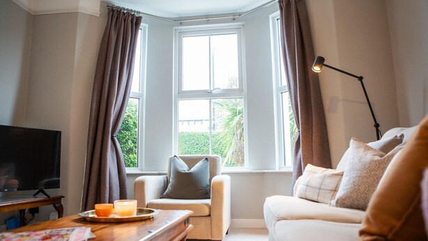 A living room with a TV, a wooden table, a sofa, and a chair. There are two windows with curtains, a lamp, and a view of greenery outside. - The Mount Street Townhouse (Harrogate)