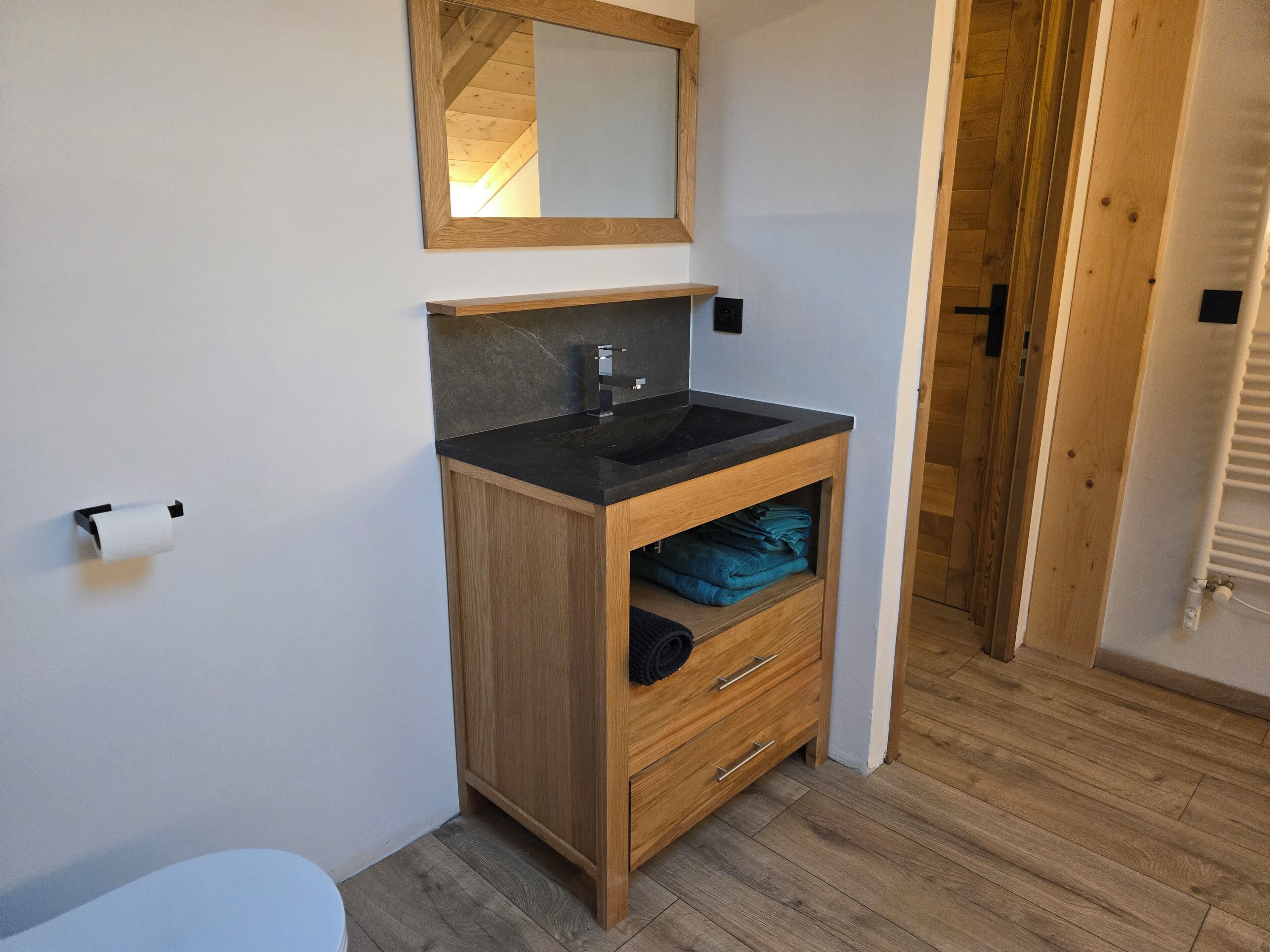 Un baño con tocador de madera, un lavabo negro y un espejo encima. Hay un dispensador de papel higiénico en la pared y un perchero con toallas.