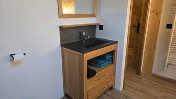 Un baño con tocador de madera, un lavabo negro y un espejo encima. Hay un dispensador de papel higiénico en la pared y un perchero con toallas.