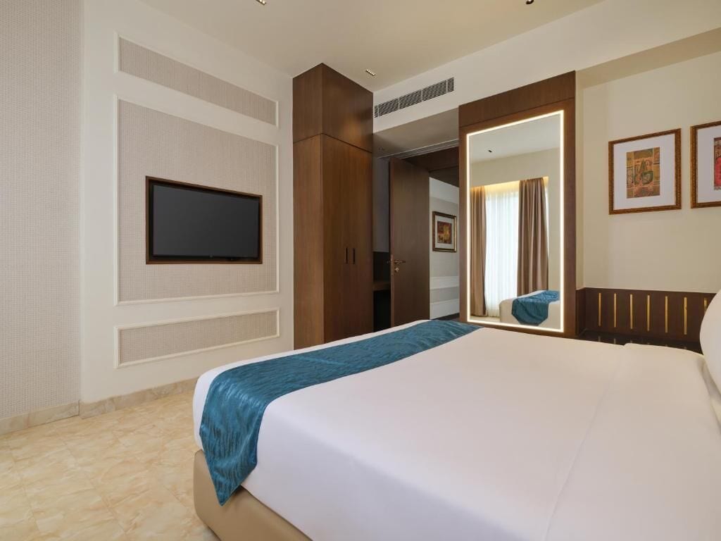 A hotel room with a large bed, a television mounted on the wall, a wardrobe, and a mirror with a light.