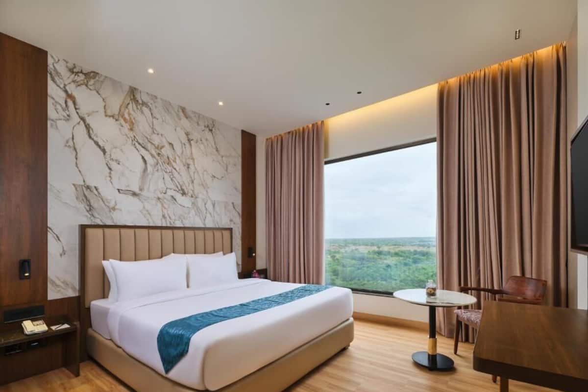 A hotel room with a large bed, a small round table, a chair, a nightstand, and a view of the outdoors.