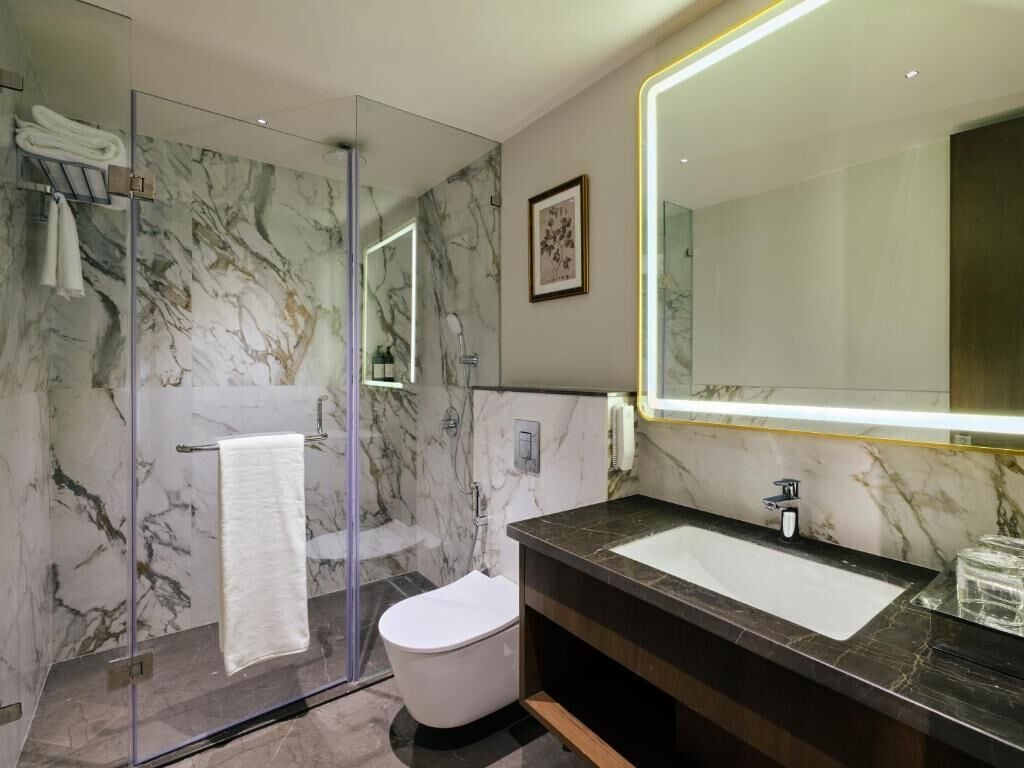 A modern bathroom with a glass shower enclosure, a white toilet, a marble-tiled wall, and a large mirror with a lighted frame.