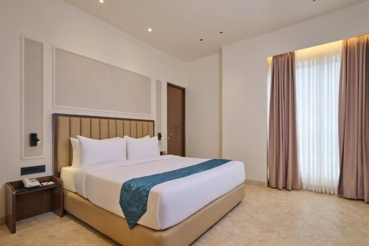A hotel room with a large bed, a bedside table, a nightstand, a wall-mounted telephone, and a window with curtains.