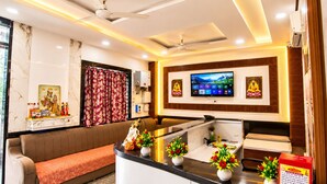 A hotel reception area with a sofa, a counter with plants, a television, and ceiling fans. - Hotel Mero Vrindavan (Vrindavan)