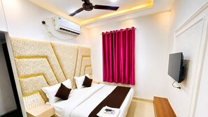 A hotel room with a bed, a bedside table, a television mounted on the wall, and a ceiling fan. - Hotel Mero Vrindavan (Vrindavan)