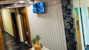 A hotel lobby with a marble reception desk, a ceiling fan, a flat-screen TV mounted on the wall, and a patterned wallpaper design. - Hotel kings villa (Ahmedabad)