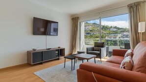 Ein modernes Wohnzimmer mit einer Couch, einem an der Wand befestigten Fernseher, einem kleinen Tisch und Blick auf eine Stadtlandschaft durch große Fenster.