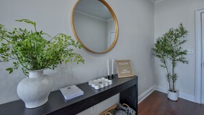 Un couloir avec une console sombre, un grand miroir ovale, deux plantes en pot et un panneau encadré.