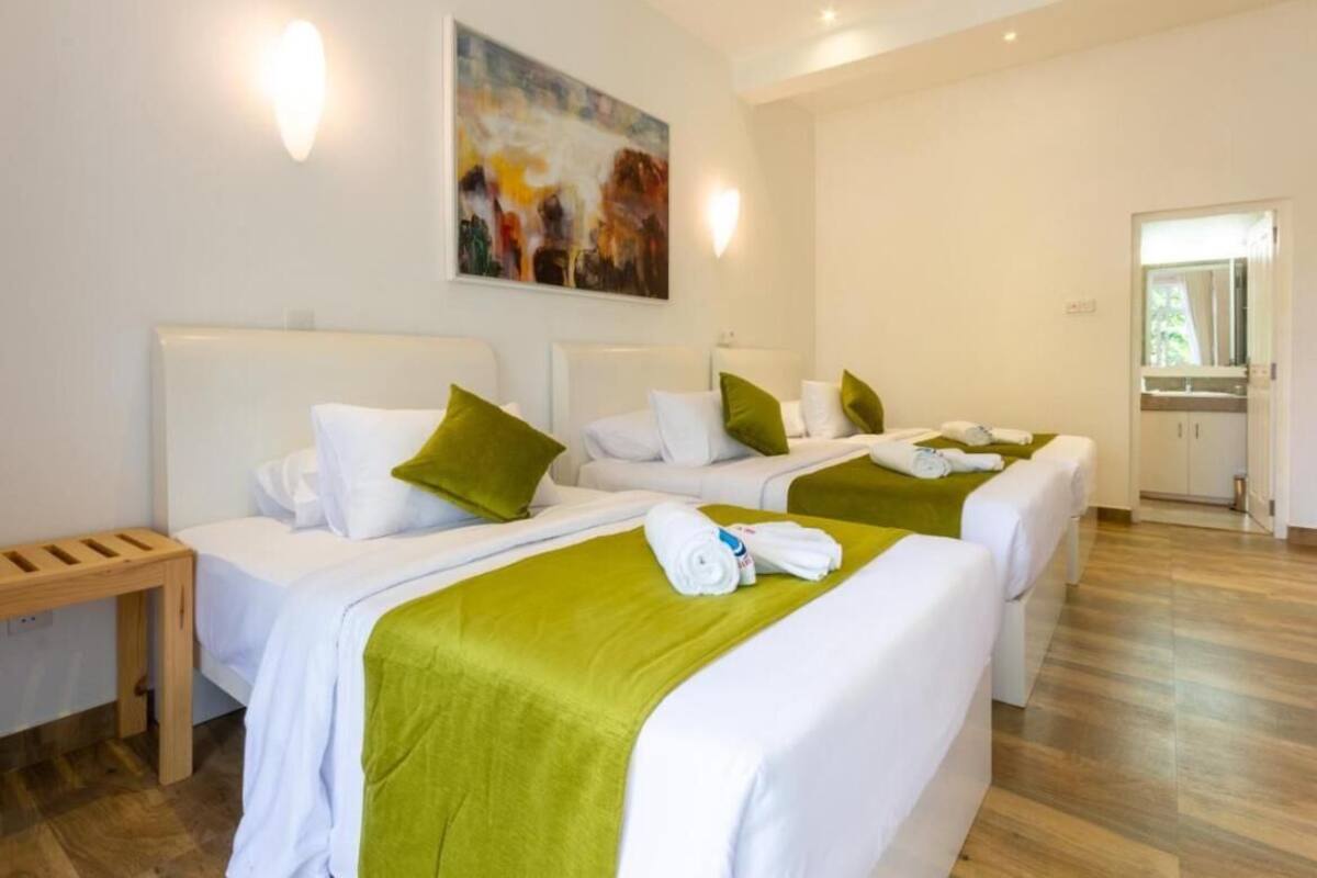 A hotel room with three beds, each with white bedding and green throw pillows. There is a wooden bench beside the beds, and a painting is visible on the wall.