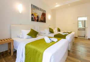 Habitación de hotel con tres camas, cada una con ropa de cama blanca y almohadones verdes. Junto a las camas hay un banco de madera, y se aprecia un cuadro en la pared.