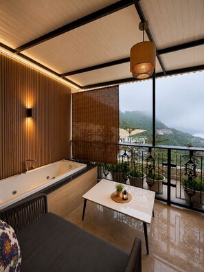 A modern balcony with a bathtub, a table with plants, and a view of a hilly landscape.