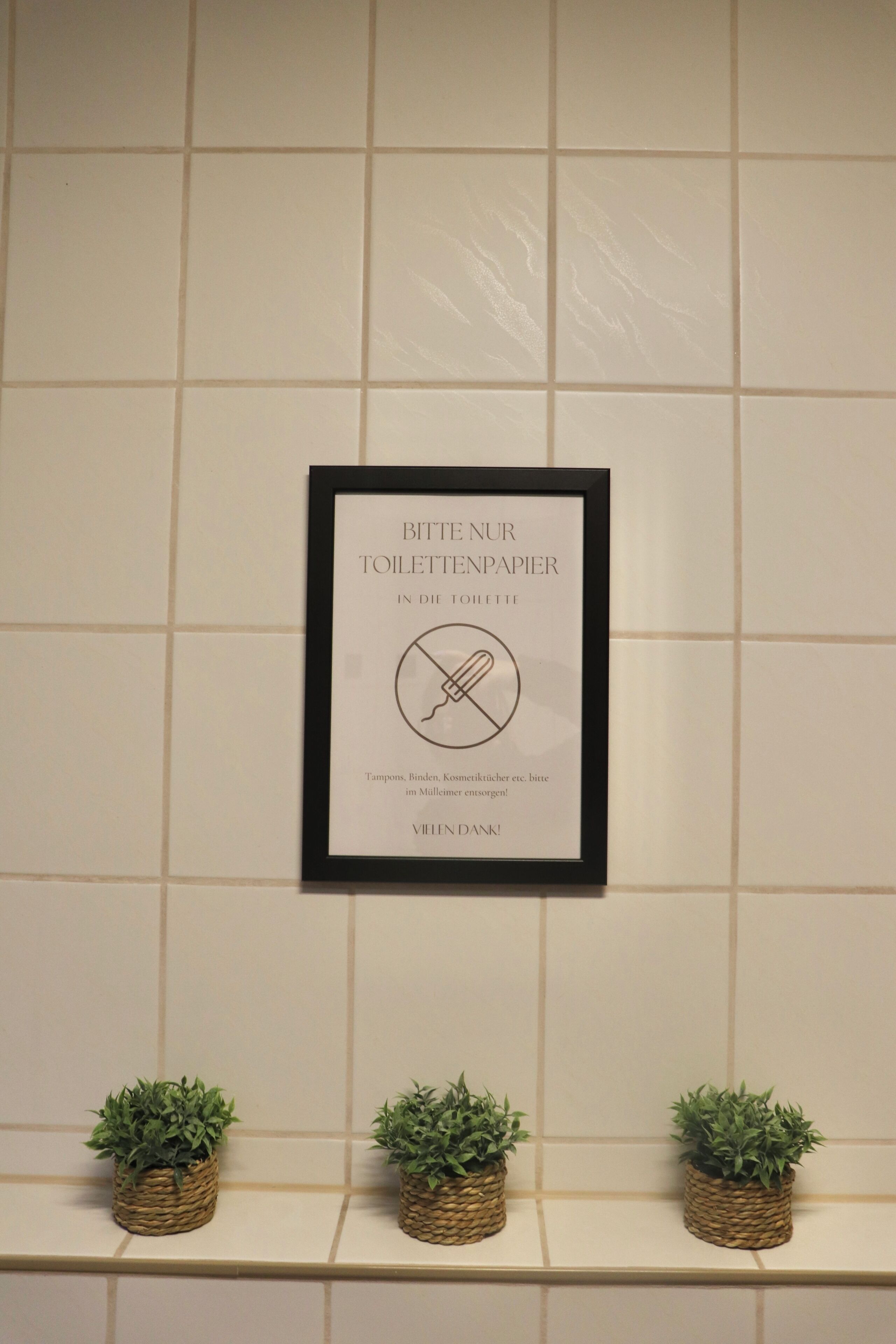 A framed sign on a tiled wall with three small potted plants below it.