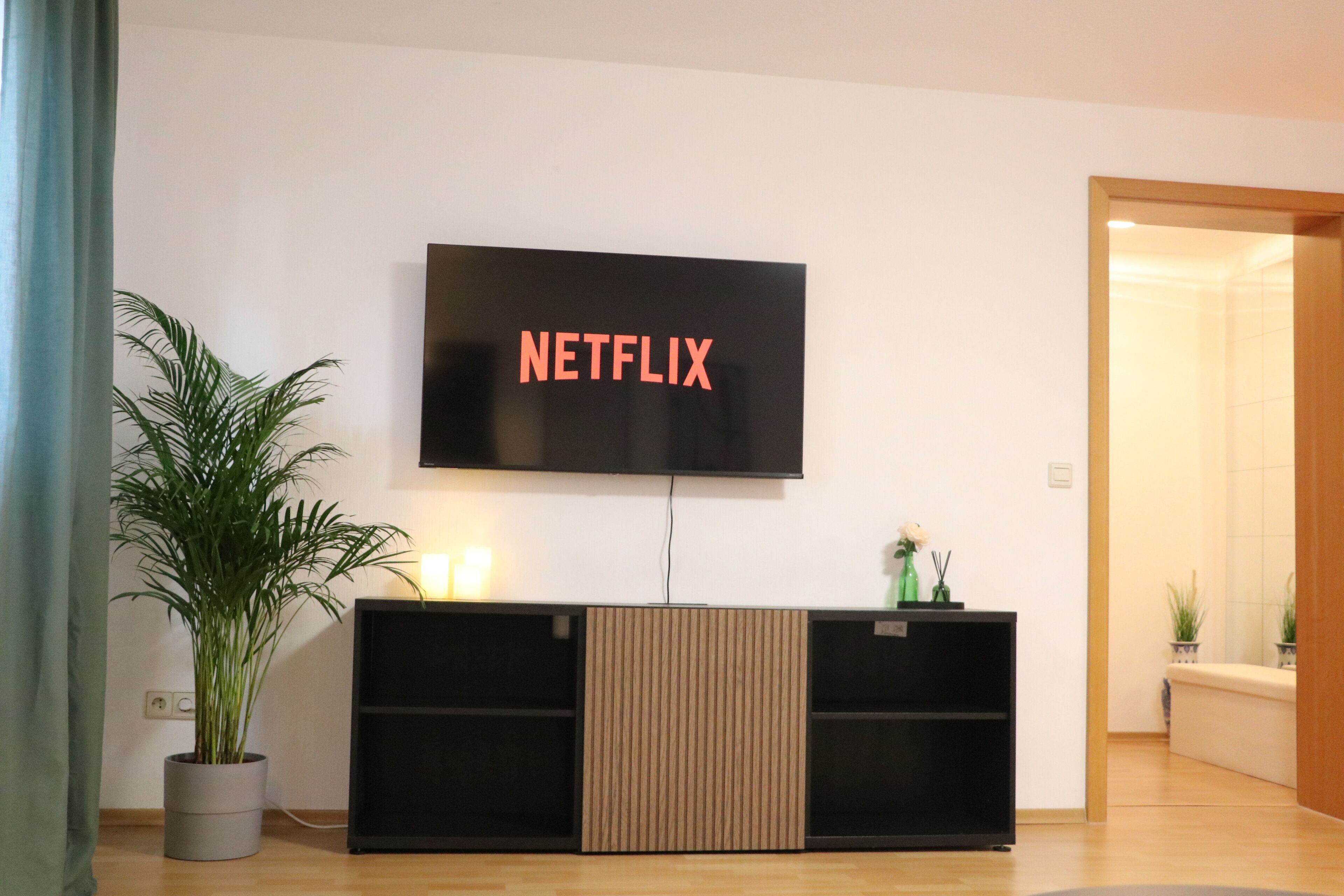 A modern living room with a flat-screen TV mounted on the wall, a black entertainment unit below, and a potted plant to the left.