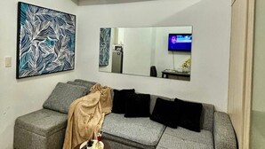 A modern living room with a grey sofa, a small round table with a plant, a large abstract painting, and a mirror reflecting the room.