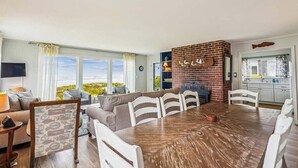 A spacious living area with a large wooden dining table, a brick accent wall, and a view of the outdoors. - Cape Escape - New Oceanfront Listing! (Arch Cape)