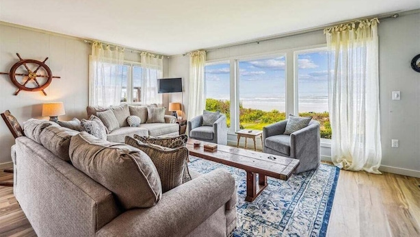 A spacious living room with a large sofa set, a coffee table, and a TV. There are large windows with curtains, a ship's wheel decoration, and a view of the sea. - Cape Escape - New Oceanfront Listing! (Arch Cape)