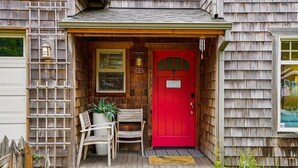 A wooden house with a red door, a porch with two chairs, and a small garden area.