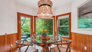 A dining area with a glass table surrounded by chairs, a wooden floor, and a large window with a view of greenery.