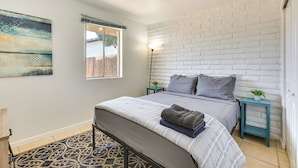 Un dormitorio con una cama, una ventana, una obra de arte colgada en la pared, una mesita de noche con una lámpara y una alfombra estampada.