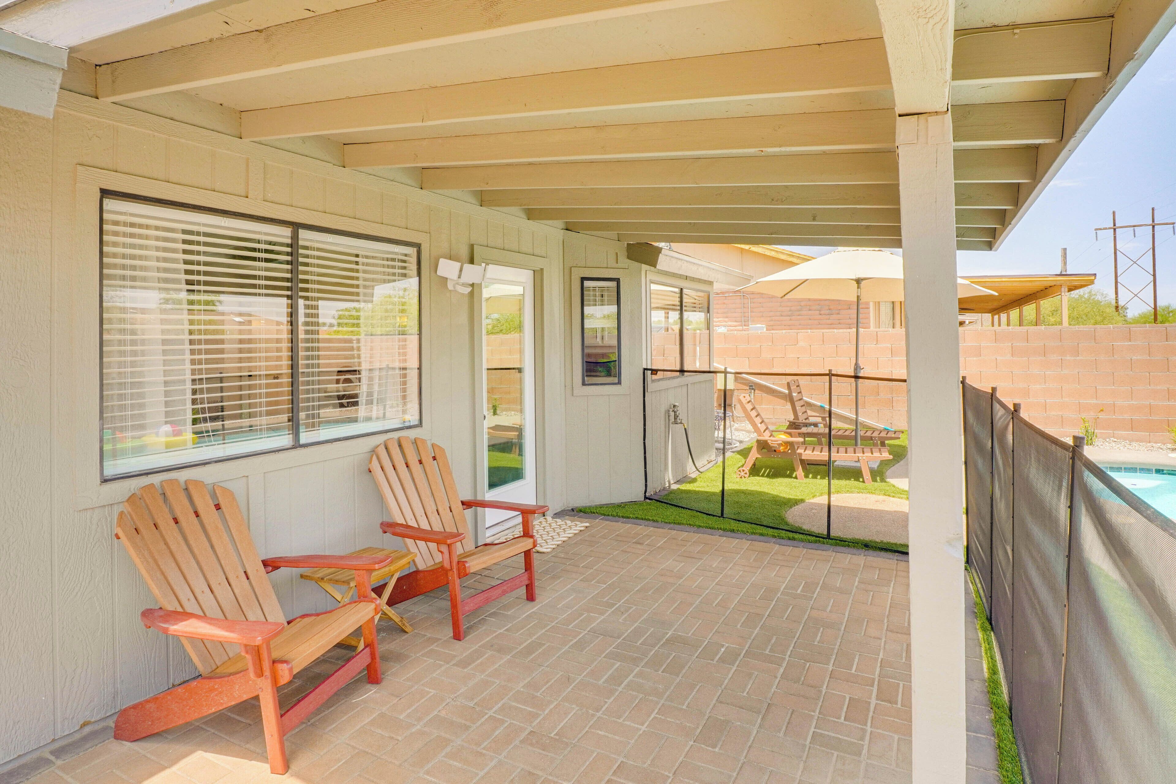 A covered patio area with two Adirondack chairs, a brick floor, and a view of a swimming pool.