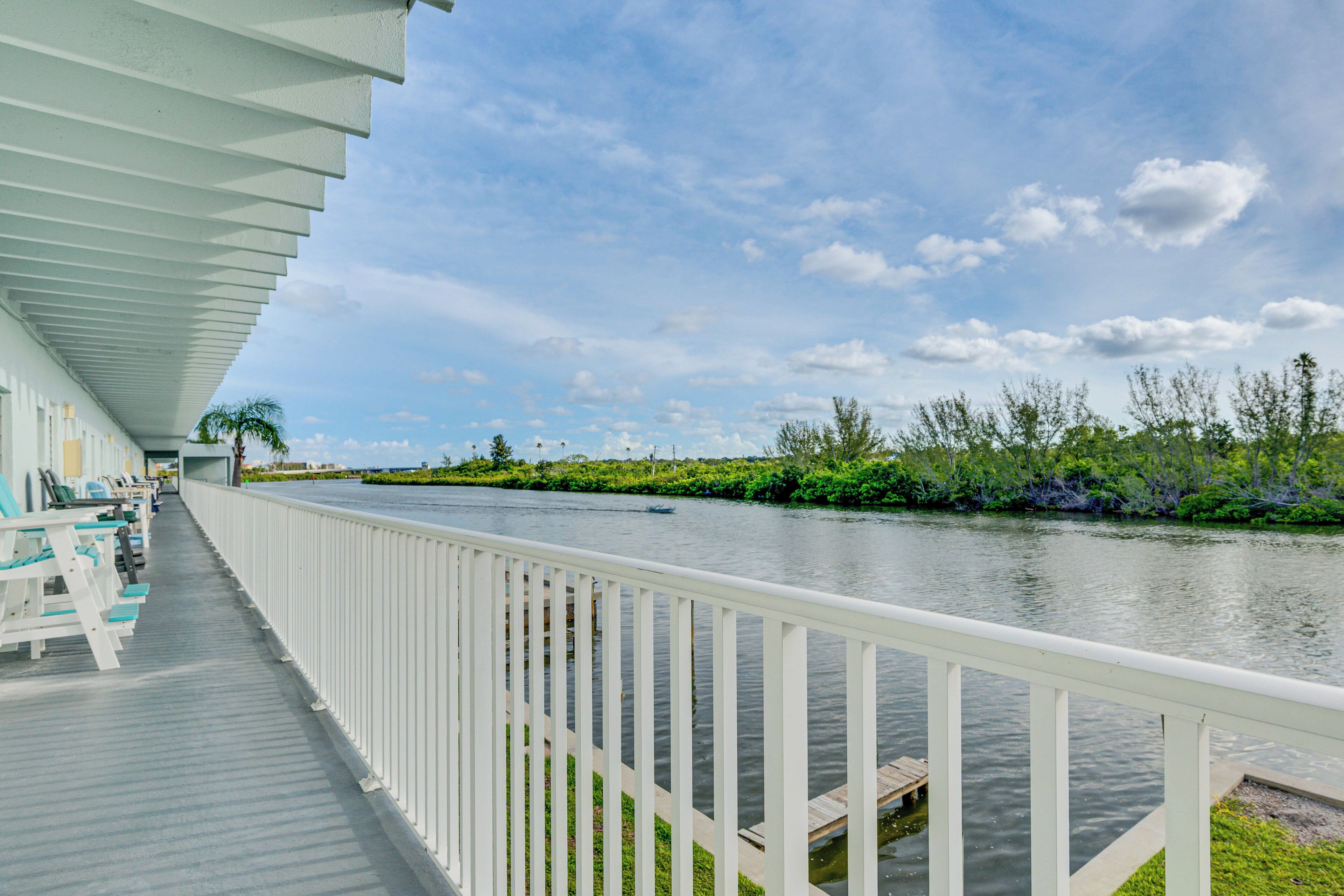 A waterfront balcony with white railings, a view of a river, and outdoor seating.