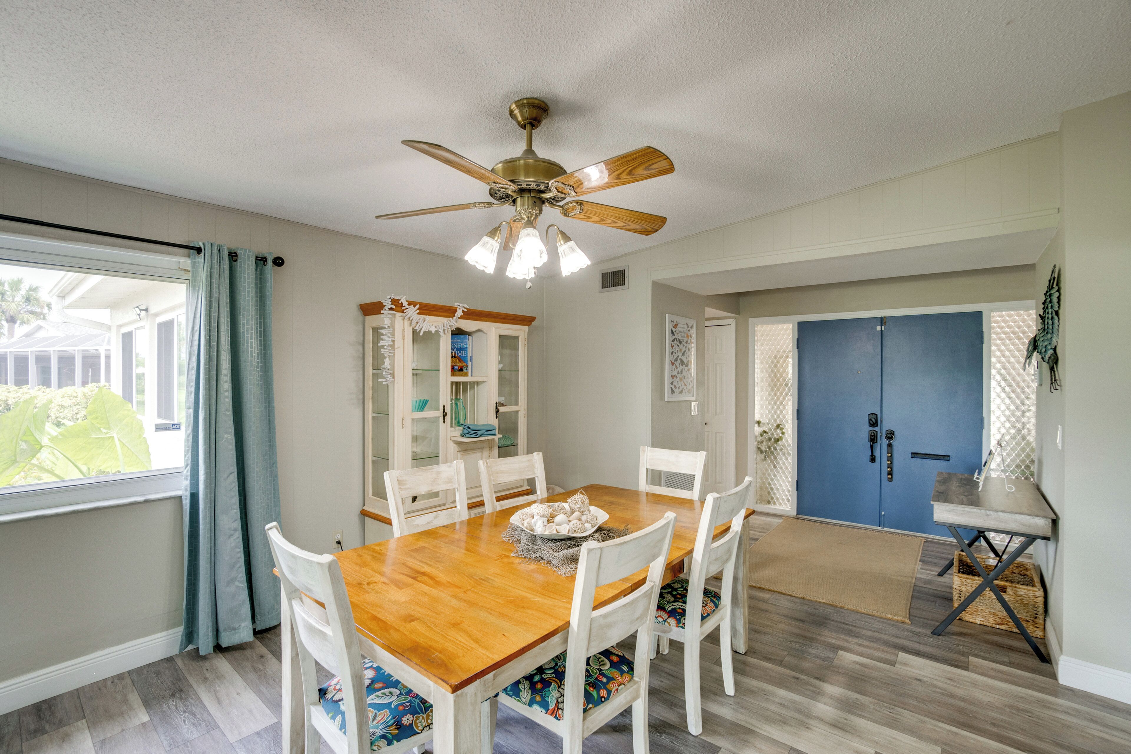 A dining area with a wooden table, white chairs, a ceiling fan, and a view of a blue door and a window with curtains.