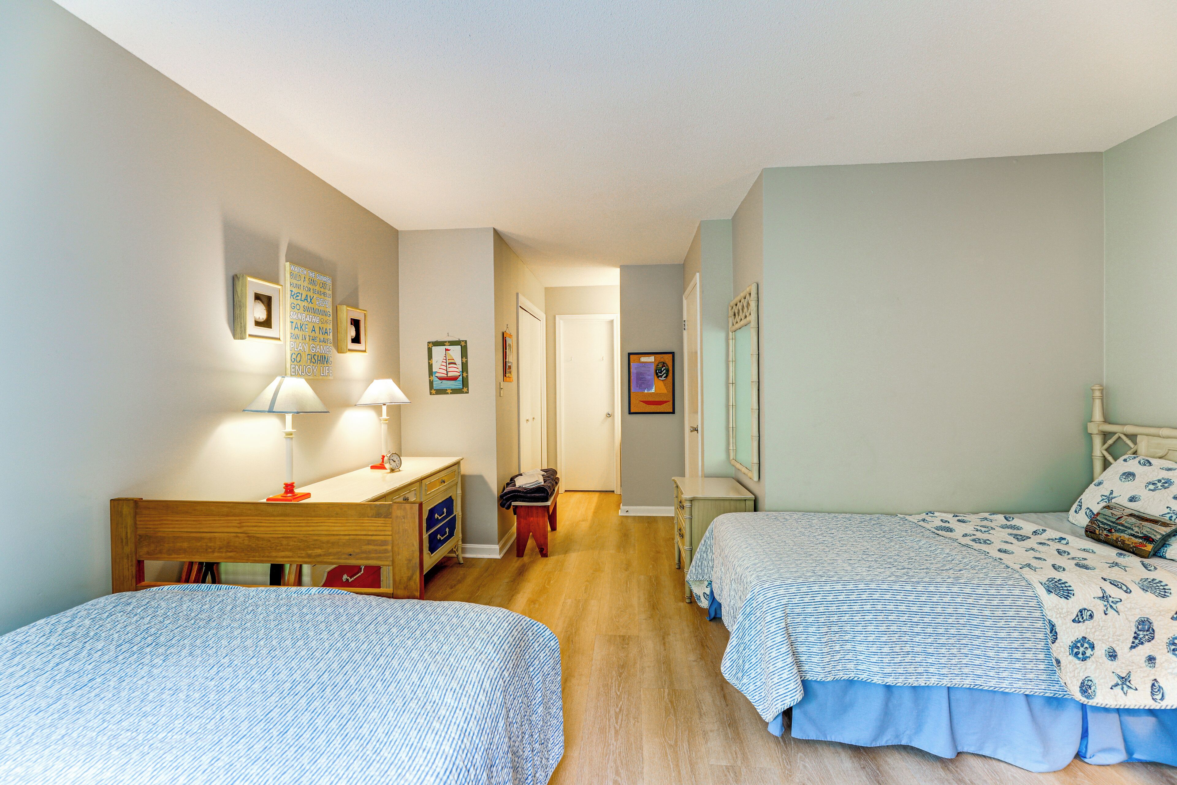A bedroom with two beds, a desk, and a chair. There are framed pictures on the walls and a door leading to another room.
